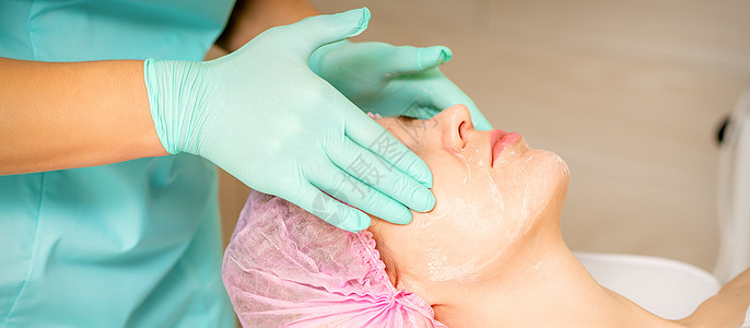 戴着手套的美容师在女性脸上涂上带去角质霜的保湿面膜 面部美容治疗 面部护理程序手指化学品化妆品去角质肤色皮肤女士剥皮凝胶按摩图片