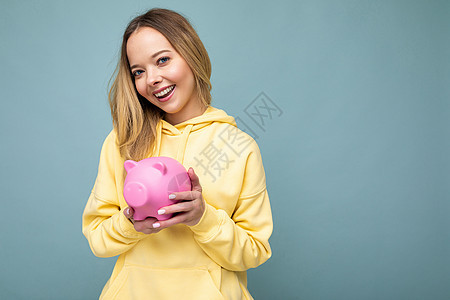快乐积极微笑的年轻美丽迷人的金发女人的肖像照片穿着时尚的黄色连帽衫与复制空间隔离在蓝色背景和持有粉红色的小猪盒与真诚的情感 存钱图片