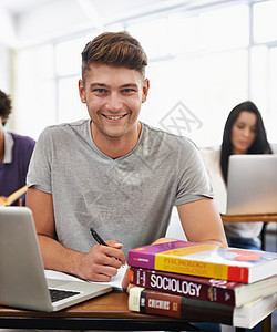 一位英俊的年轻学生坐在书桌旁 带着教科书坐着呢学者图书技术男性学校学习成人微笑学习者电脑图片