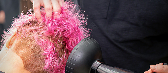 在美发沙龙 一位年轻的白人女性用黑色吹风机用刷子吹干粉红色的短鲍勃发型 特写闲暇烘干头发商业治疗横幅工作理发师爆头烘干机图片