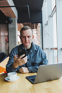 白天 穿着休闲装 面带微笑的男性企业家坐在桌边 拿着笔记本电脑 在带大窗户的现代工作区用智能手机聊天桌子职场自由职业者咖啡互联网图片