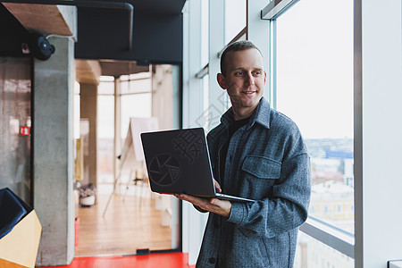 一个聪明聪明的男性经理的肖像 他拿着一台笔记本电脑在办公室进行研究 一个穿着优雅衣服的商务人士在触摸板上玩 站在办公室的大窗边职图片
