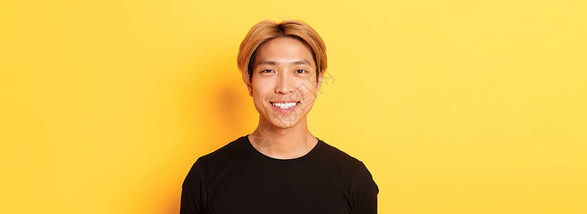 身穿黑色 T 恤的金发亚洲帅哥的特写 对着镜头笑得很开心 站在黄色背景上情感头发学生广告快乐理发成人发型促销购物图片