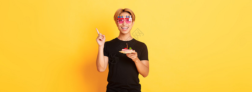 快乐 帅气的亚洲生日男 戴着滑稽的太阳镜 拿着带蜡烛的生日蛋糕 手指右上角指着你的标志 黄色背景职业学生男性成人广告促销头发男朋图片