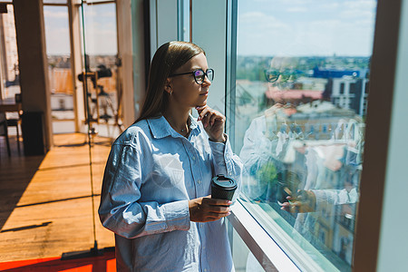 一位戴眼镜的年轻女性自由职业者端着咖啡站在一扇大窗户旁 看着这座城市 一个穿着衬衫和短裤的女孩站在窗边的咖啡馆里女士手机城市办公图片