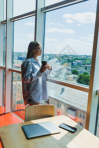 一位戴眼镜的年轻女性自由职业者端着咖啡站在一扇大窗户旁 看着这座城市 一个穿着衬衫和短裤的女孩站在窗边的咖啡馆里办公室技术女士人图片