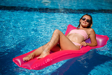 穿着粉红色比基尼泳衣的女人漂浮在可充气的粉色床垫上 Spf和防晒霜是被涂过的太阳女性女孩魅力假期游泳派对微笑海滩泳装图片