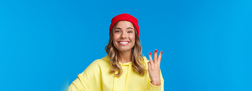 穿着红豆和黄连帽衫的英俊caucasians女性 展示第3号 微笑 秩序或保留 自豪和高兴 蓝背景学生教育假期工作室大学青年成人背景图片