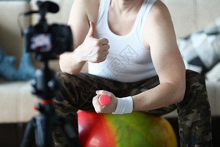 运动员男子举起拇指抬起哑铃 当着摄像机的面坐在家里的球上图片