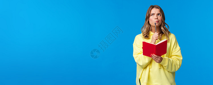 深思熟虑的金发欧洲女性制定计划 用笔触摸嘴唇 在思考做笔记时抬头思考 拿着红色笔记本 站在蓝色背景家庭作业大学微笑成人学生日记写图片