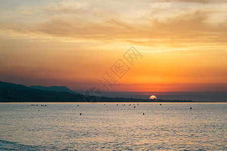 在海洋的五颜六色的橙色早晨日出 冲浪者在平静海浪中的剪影与多云的天空相映成趣 涂成亮橙色 第一缕阳光反射出水面图片