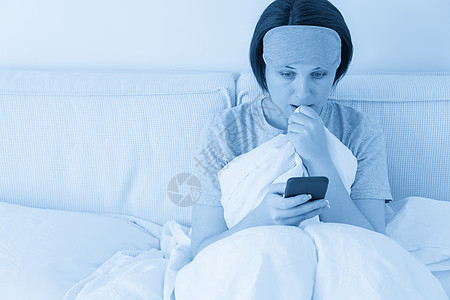 戴着睡眠面具的女人坐在铺着羽绒被的床上 用智能手机发短信女孩蓝色卧室房子技术房间女士互联网黑发说谎图片