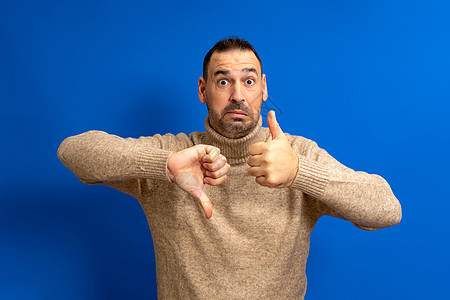 佩戴高领毛衣的胡子西班牙男子举起拇指和拇指 以怀疑和困惑的方式与蓝色背景隔绝 (掌声)图片