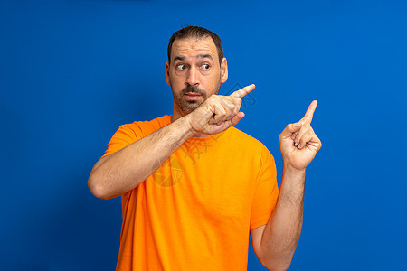 身穿橙色T恤的胡子手部男子指向侧面 在蓝工作室背景上用索引手指隔离图片