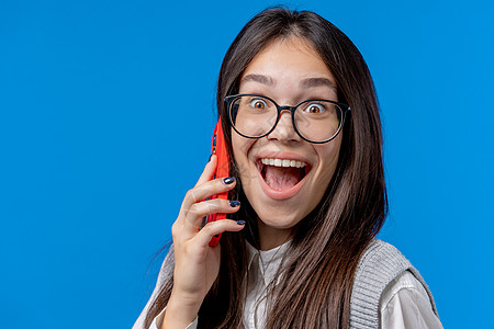 令人吃惊的亚洲学生青少年与朋友通过电话交谈 开个好玩笑 年轻女孩张着嘴说蓝底话 打过电话图片