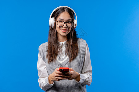 漂亮的亚洲少年在智能手机上听音乐和上网 在蓝色背景上带着微笑和喜悦的女孩 科技 成功 幸福 社交网络概念图片