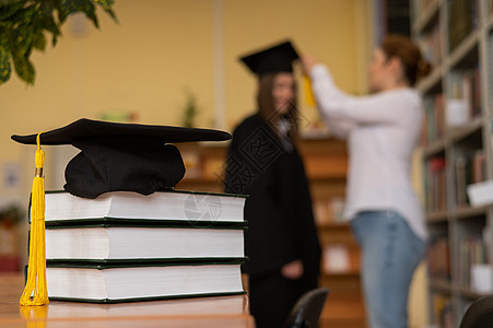 在图书馆的一堆书上加盖了毕业帽 教师和背景中的女青年毕业生都完成了毕业封套学习智慧成就考试班级仪式校园教科书大学老师图片