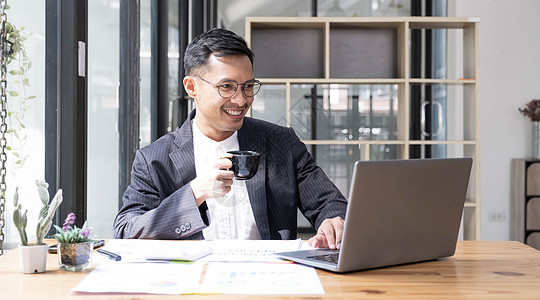 年轻的亚洲商务人士穿着西装 拿着热咖啡杯 看着办公室的笔记本电脑 在光线充足的工作场所使用笔记本电脑的西装男士管理人员男性技术套图片