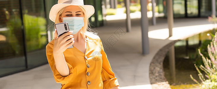一位戴着医用口罩的漂亮年轻白人女子提着手提箱走在街上 在冠状病毒流行高峰期 一个背着行李的女孩独自旅行 空城商业感染学生发烧车轮图片
