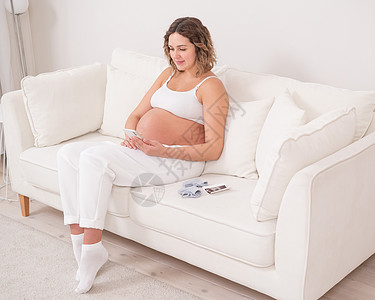 孕妇坐在白色沙发上 有智能手机 胎儿超声波检查家庭肚子享受妇科腹部照片生育力电话母亲新生活图片