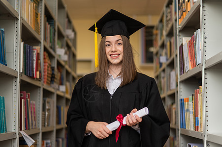 穿着毕业礼服的快乐年轻女子 在图书馆持有文凭教育微笑大学女孩证书女性庆典书架学生地幔图片