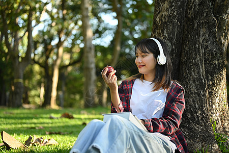 快乐的年轻女士 在大树下吃苹果 绿草上 阳光明媚美丽的花园背景图片