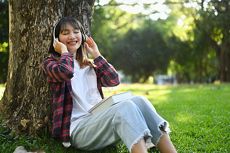 快乐的亚洲女人喜欢听耳机音乐 在阳光明媚的美丽白天坐在绿草树下的青草下图片