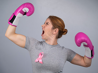 胸口有粉色丝带的妇女用灰色背景的粉色拳击手套举起手来 战胜乳腺癌微笑女性精力力量斗争冒充卫生女孩保健拳头图片