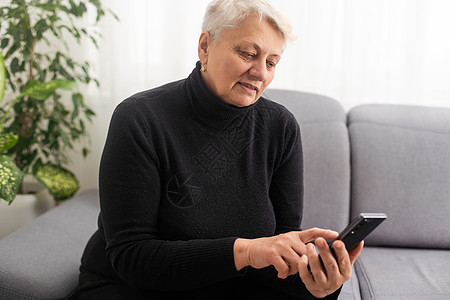 快乐成熟的 60 多岁老妇人拿着智能手机使用手机应用程序进行视频通话 一边看有趣的视频一边笑 坐在家里的沙发上兴奋地在手机上赢得图片