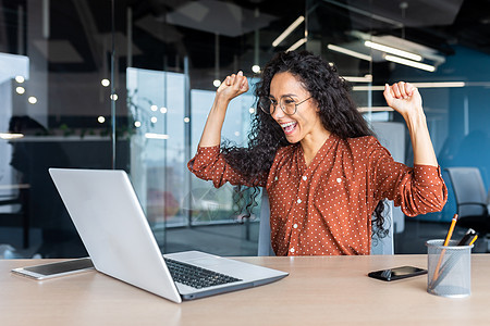西班牙裔女商务人士庆祝胜利成功 办公室里卷发的员工阅读好消息 在办公室工作时使用笔记本电脑举起手 做出快乐的胜利手势成就优胜者窗图片