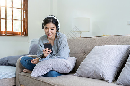 亚洲女人用手机和耳机听音乐 平静的家庭周末 坐在沙发上放松 享受最喜欢的曲调 流行音乐 在客厅使用现代无线耳机和小工具闲暇播客成图片