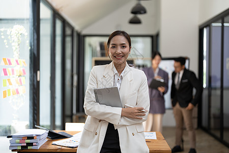 身着正式商务套装的年轻迷人亚洲女办公室职员的画像在办公室对着镜头微笑 背景是模糊的同事在办公室工作职业管理人员经理女性工作室员工图片