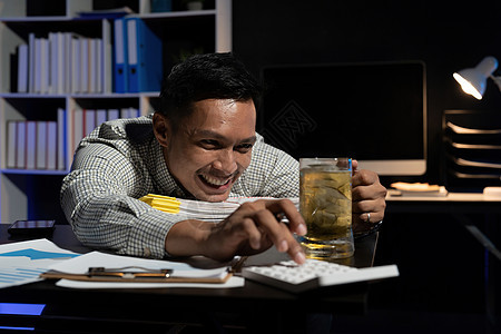 亚洲男子在晚上办公室使用计算器时加班喝酒喝啤酒 而他却在办公室工作图片