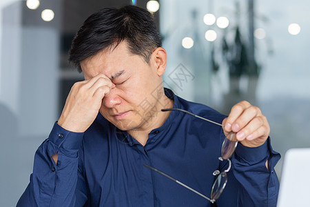 一个疲累的亚洲年轻男人坐在办公室里 戴眼镜 用手闭上眼 用手闭上眼睛图片