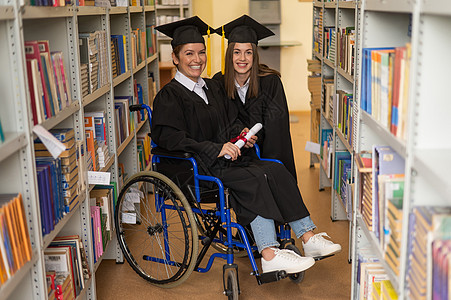 快乐的年轻女性和穿着研究生礼服坐在轮椅上的妇女 在图书馆拥有文凭 包容性教育自由流苏学校学生学士证书仪式学习地幔多样性图片