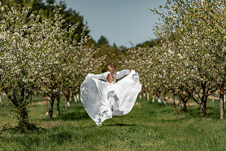 穿着白裙子的女人穿过开花的樱桃园 长裙飞向两侧 新娘一生都欢欣鼓舞 笑声晴天公园女士乐趣树木自由婚礼果园樱花跑步图片