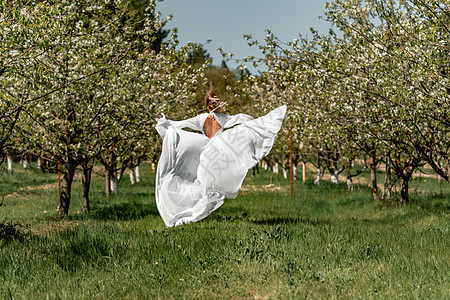 穿着白裙子的女人穿过开花的樱桃园 长裙飞向两侧 新娘一生都欢欣鼓舞 笑声晴天乐趣场地自由女士果园跑步公园树木头发图片