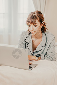 自由职业女性穿着睡衣 躺在床上 在笔记本电脑 电脑上工作 在互联网上学习 上网冲浪 放松 放松 在卧室 客厅 家里 酒店房间里度图片