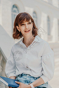 一个身穿白衬衫和蓝色牛仔裙的女人坐在矮墙上 在建筑物的背景下 手里拿着一本蓝色的笔记本 夏天在城市里散步 旅行嘴唇黑发女性办公室图片