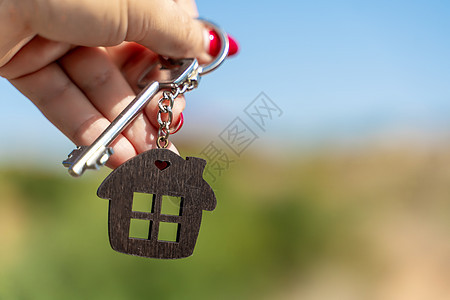 在绿树和天空的背景下 女人手里拿着房子形状的钥匙链的钥匙 购买房屋 公寓 房地产 年轻家庭 抵押贷款 出租房屋的概念房间商业钥匙图片
