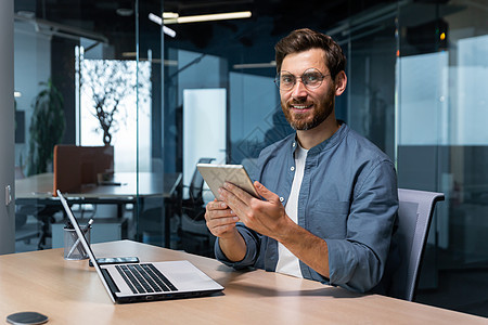现代办公室里一位商务人士的肖像 一位男士使用平板电脑 坐在办公桌前 微笑着 穿着休闲衬衫和眼镜看着镜头图片
