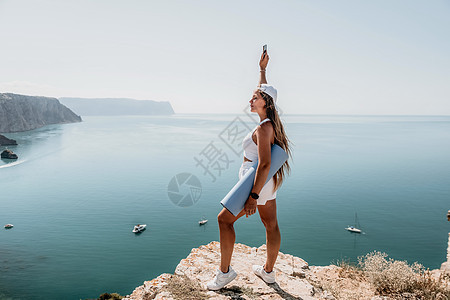 在平静的蔚蓝大海上用瑜伽垫的女人 漂亮的中年妇女留着长头发 在海边有蓝色瑜伽垫的健身教练 女性健身瑜伽常规概念 健康的生活方式教图片