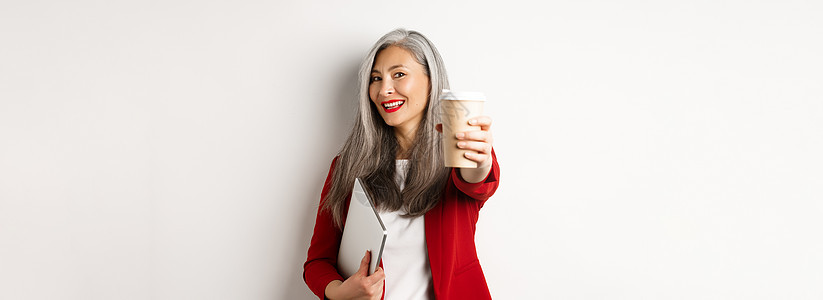 愉快的亚洲女性经理 给你一杯咖啡和微笑 用笔记本电脑站在手边 白色背景 笑声人士套装杯子商务自由职业者企业家电子商务外套工作工人图片