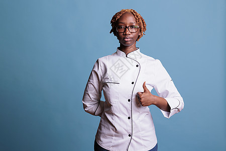 严肃的非洲裔美国女厨师站着竖起大拇指看着相机 穿着制服的专业女厨师在蓝色背景下竖起大拇指表示赞同图片