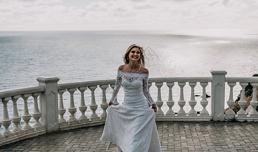 浪漫的新娘 穿着白色婚纱的金发女孩 肩部敞开 在以大海和岩石为背景的开放式露台上摆姿势 时尚的年轻女子站在露台上 在日落时望着大图片