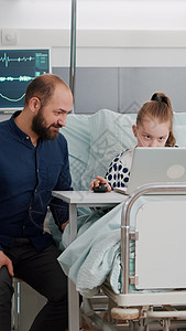 父亲坐在生病的女儿旁边 同时使用笔记本电脑在网上玩卡通视频游戏父母病房病人药品治疗临床微笑诊断互联网卫生图片