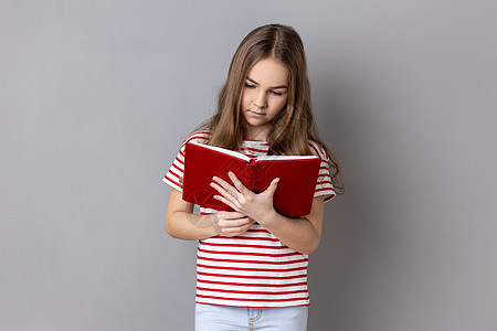 穿着带条纹T恤衫的注意力集中的小女孩持有书籍 阅读 并被有趣的阴谋所吸收图片