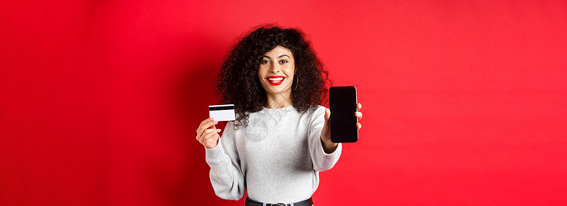 展示塑料信用卡和空智能手机屏幕的快乐漂亮女孩 展示账户或应用程序 站在红色背景上工作室购物电话女性广告技术黑发生活卷曲情感图片