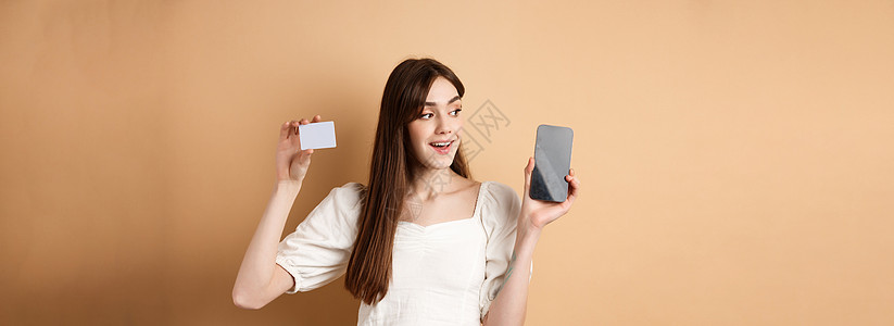 看看这个 微笑的快乐年轻女性展示塑料信用卡和空手机屏幕 演示申请 站在蜜蜂背景上站立着米色背景技术学生女孩金融假期购物电子商务女图片