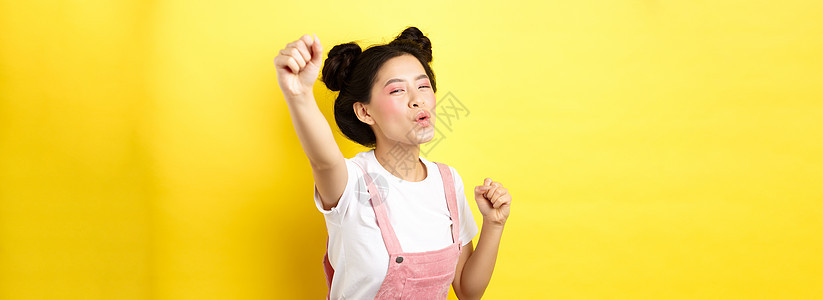 兴奋的亚洲女孩看着积极进取 举起手高呼 欢喜地欢乐 站在黄色背景上快乐头发发型美丽胜利青少年口红假期嘴唇化妆品魅力背景图片